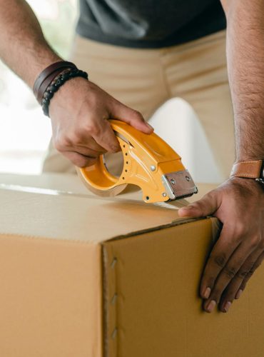 Crop faceless young male with wristwatch using adhesive tape while preparing cardboard box for transportation